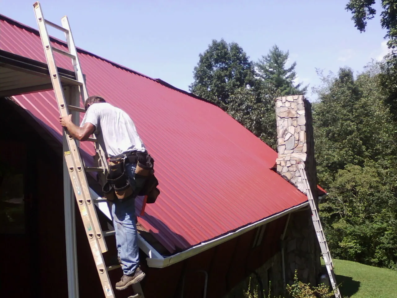 Roofer on a ladder beside a red metal roof and stone chimney — Soffit & Fascia Repair by Roofing Brothers in Blue Diamond